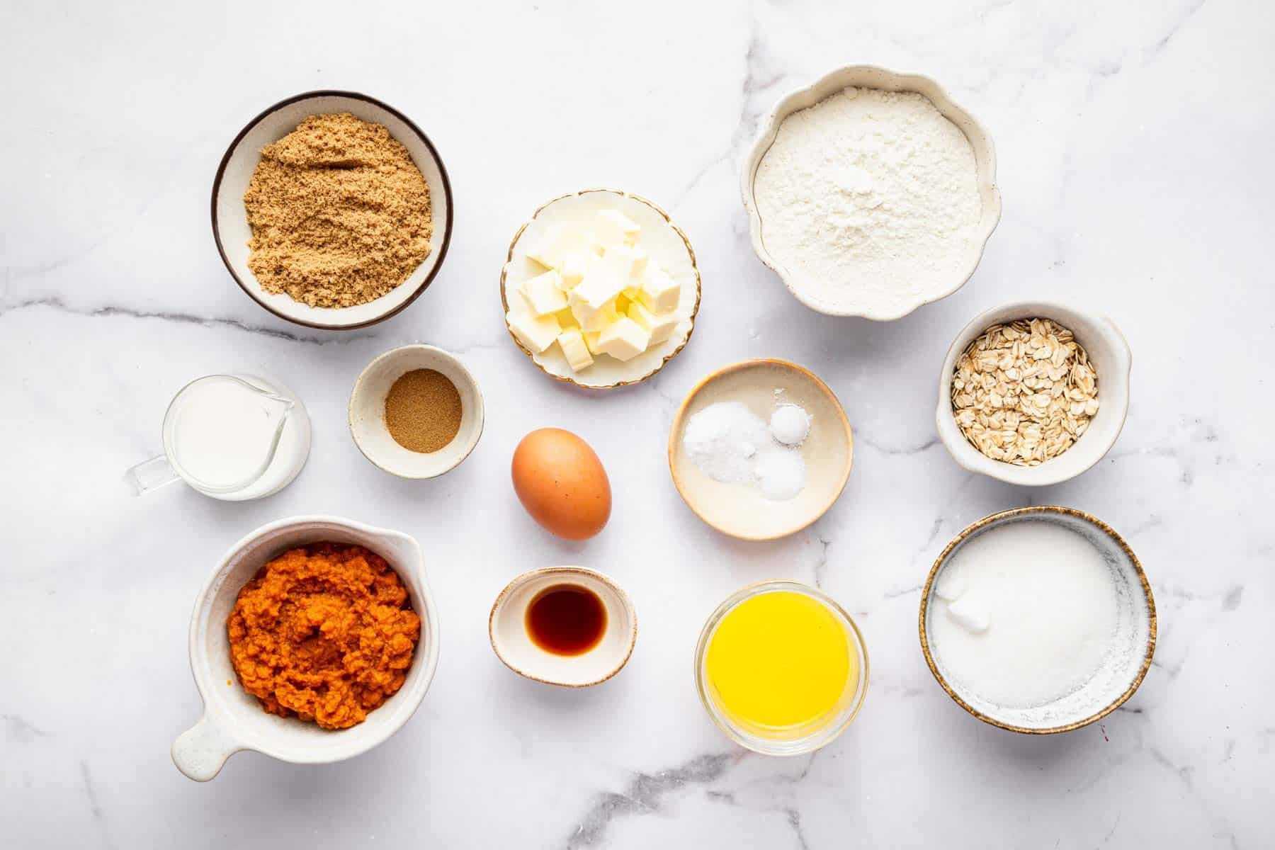 Bowl of oats, brown sugar, diced butter, egg and vanilla on marble counter.