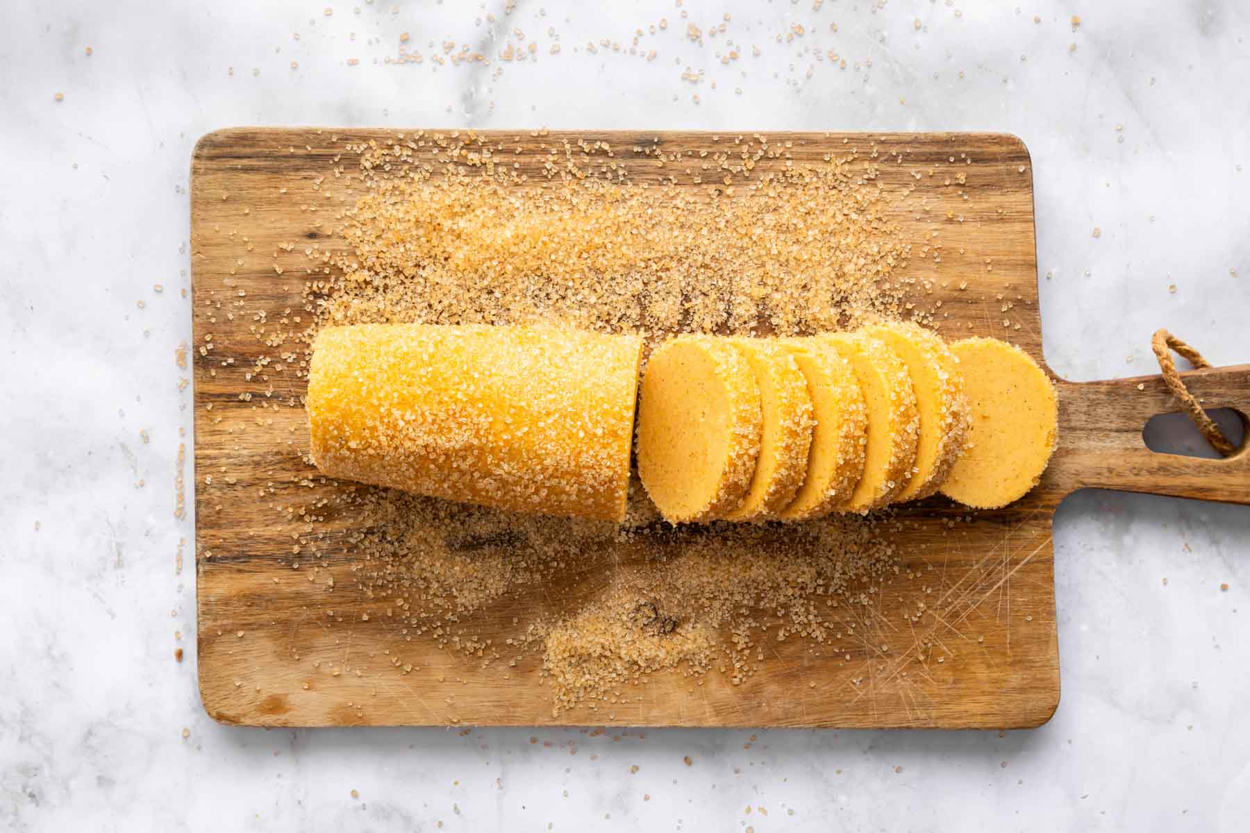 Log of orange dough rolled in coarse sugar on wooden cutting board.