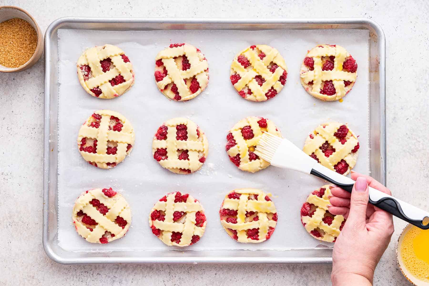 A hand brushing egg wash on fruit pastries on sheet pan.