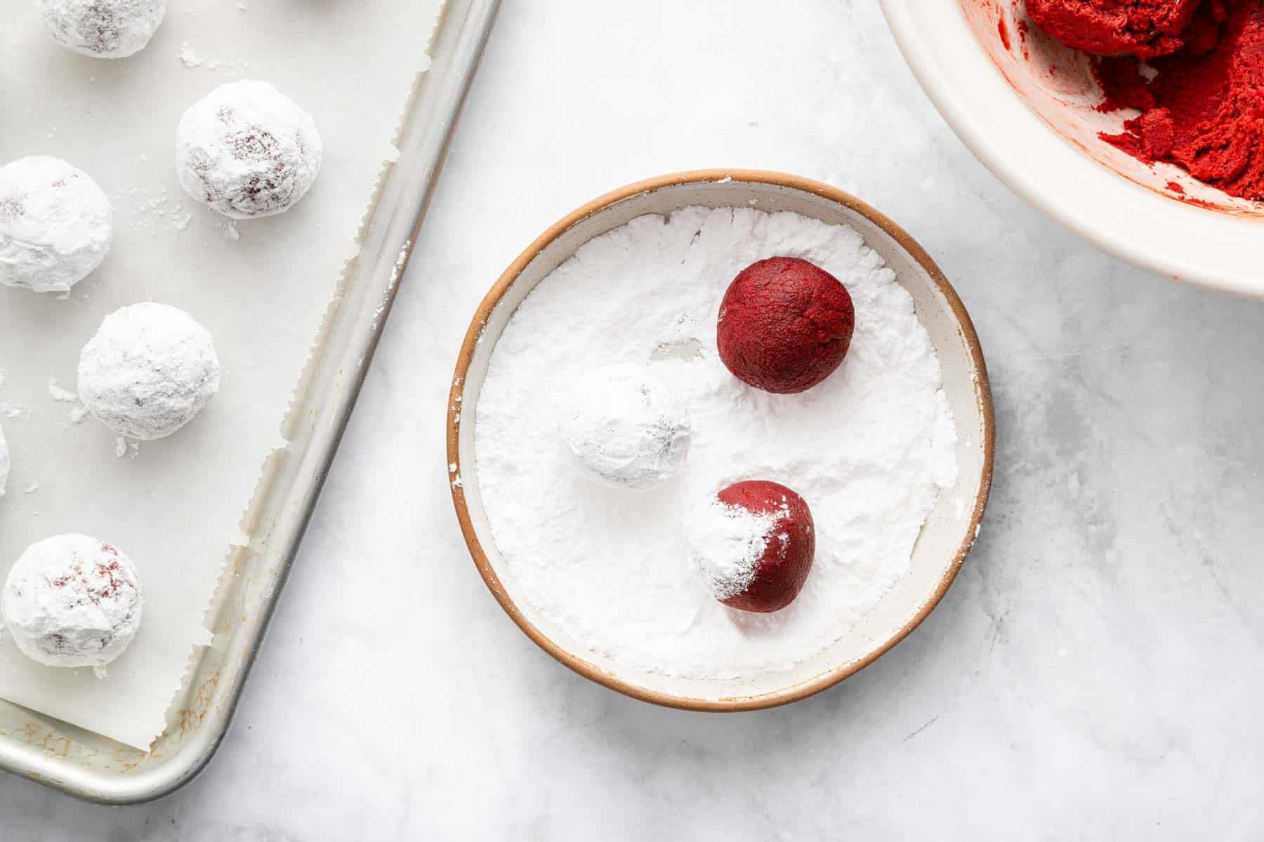 Three red dough balls being rolled in powdered sugar.