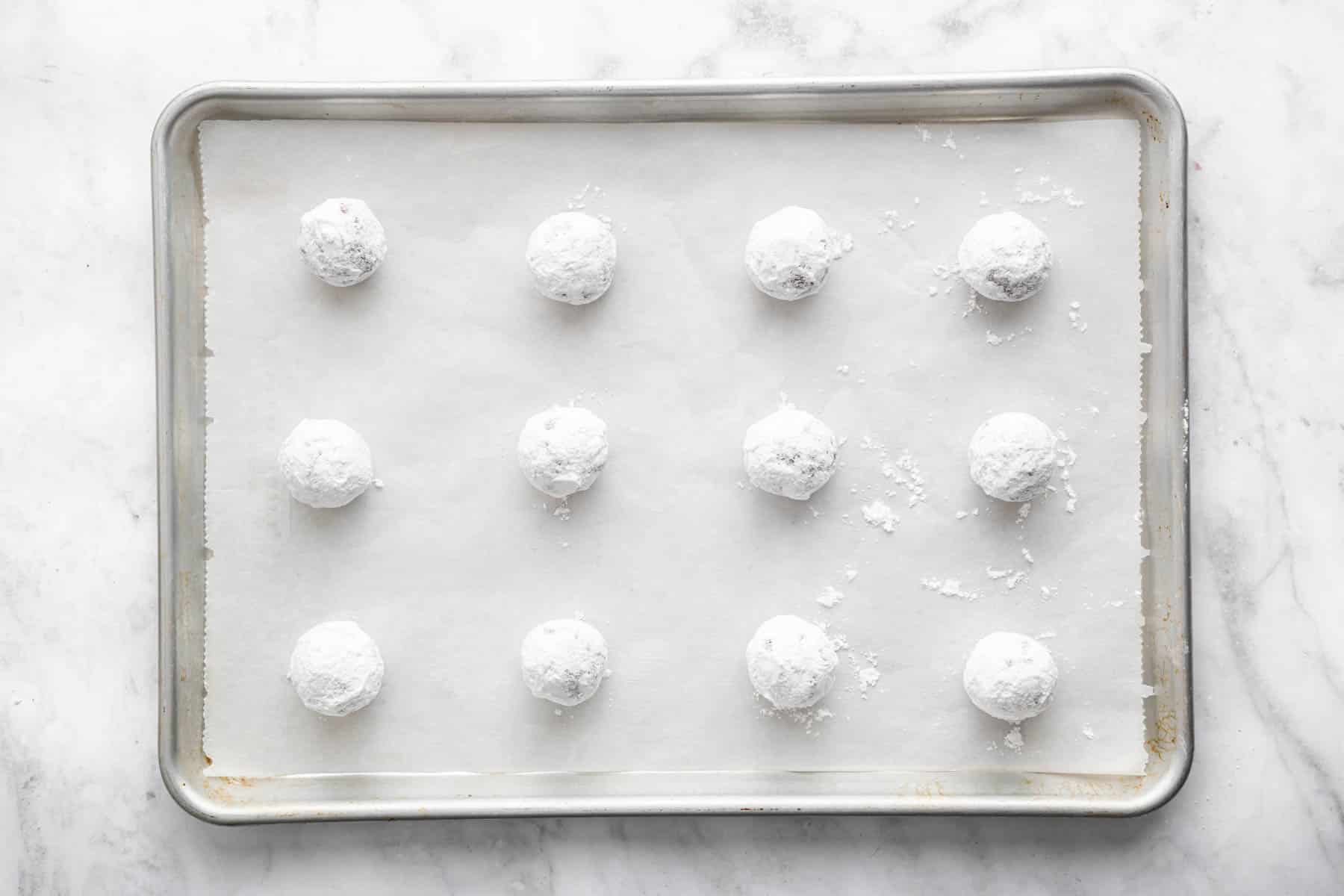 Twelve raw powdered sugar covered dough balls on a baking sheet.