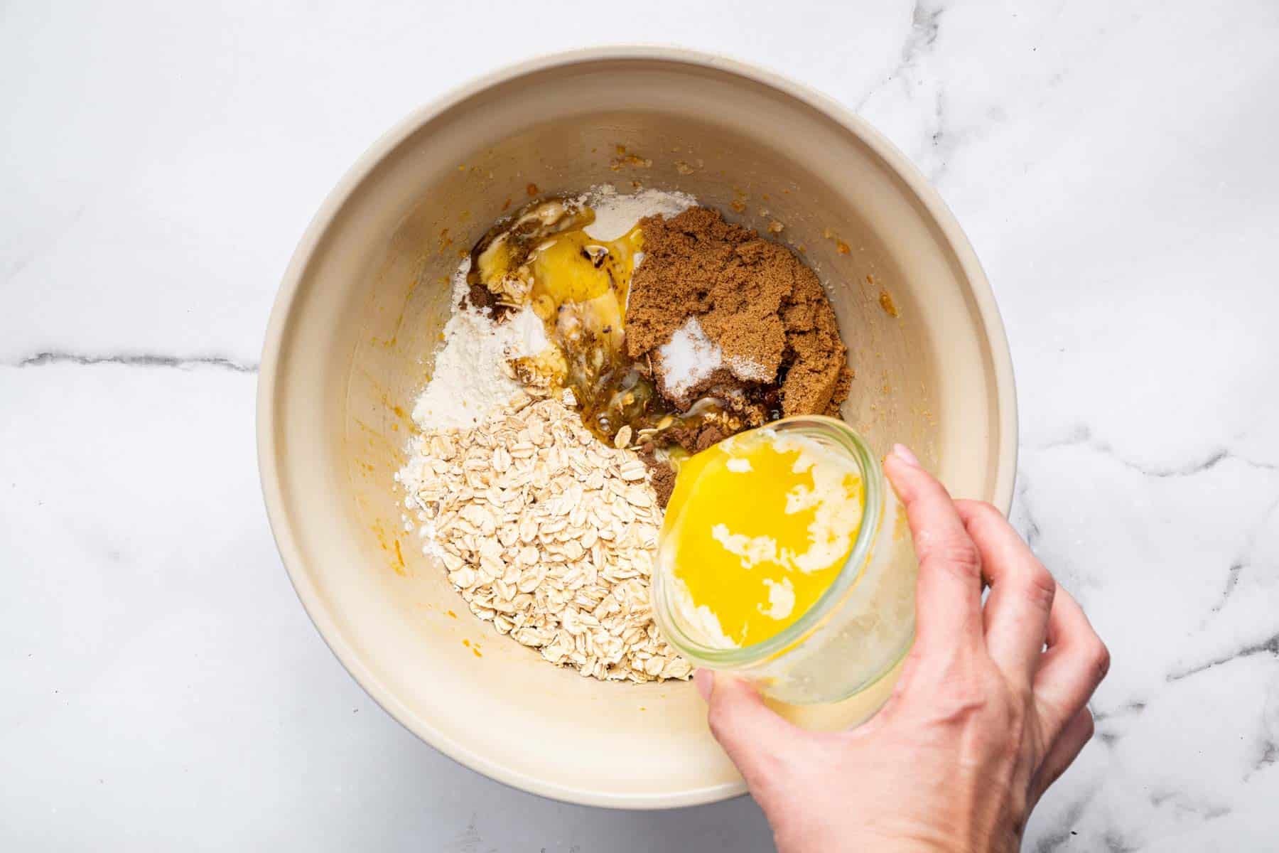 Hand pouring melted butter into a bowl with oats and brown sugar.