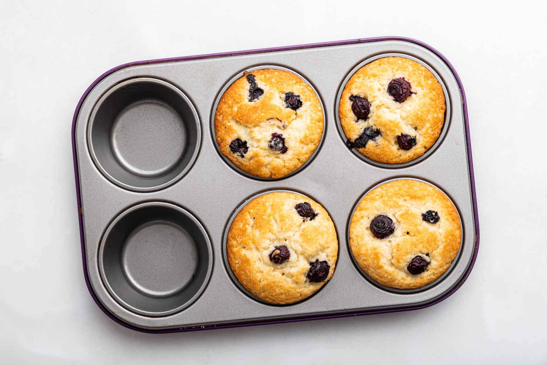 Four blueberry muffins, freshly baked in a muffin pan with 6 cups.