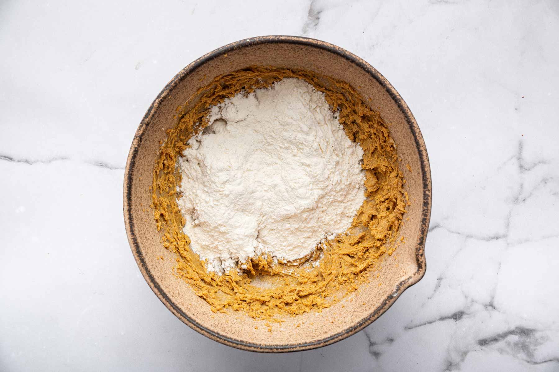 White flour in brown bowl over tan liquid dessert batter.