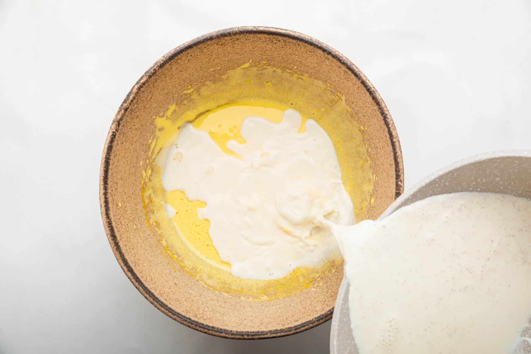 Pouring milk over yellow batter in brown bowl.
