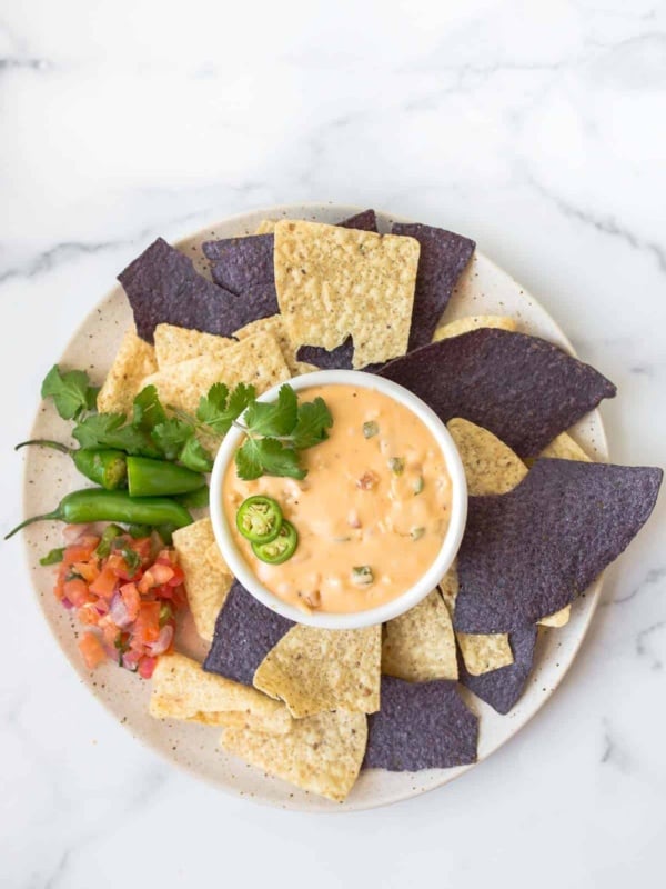 Bowl of queso with multi color tortilla chips surrounding it on plate.
