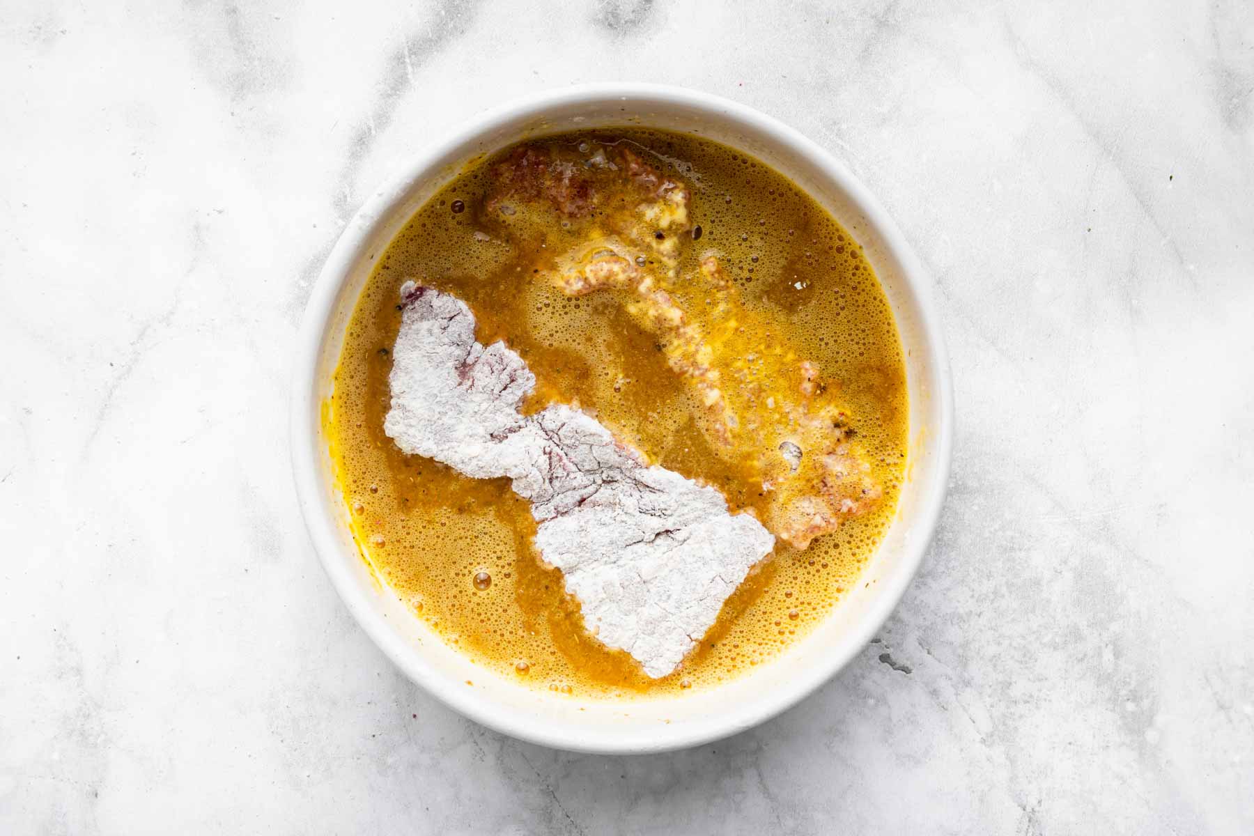 Flour coated piece of meat in bowl with yellow batter.