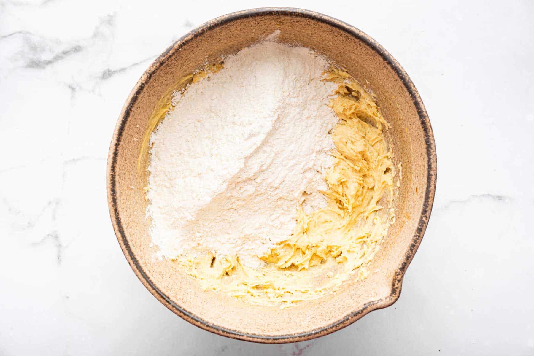 White flour on top of light yellow batter in brown bowl.