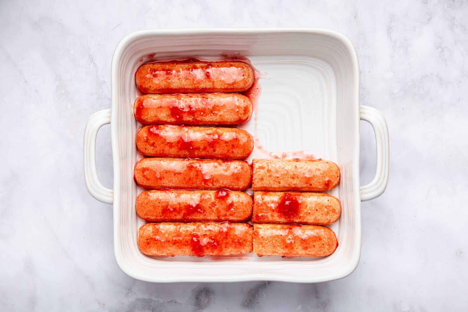 Red ladyfingers dipped and lined up in the bottom of a square pan.