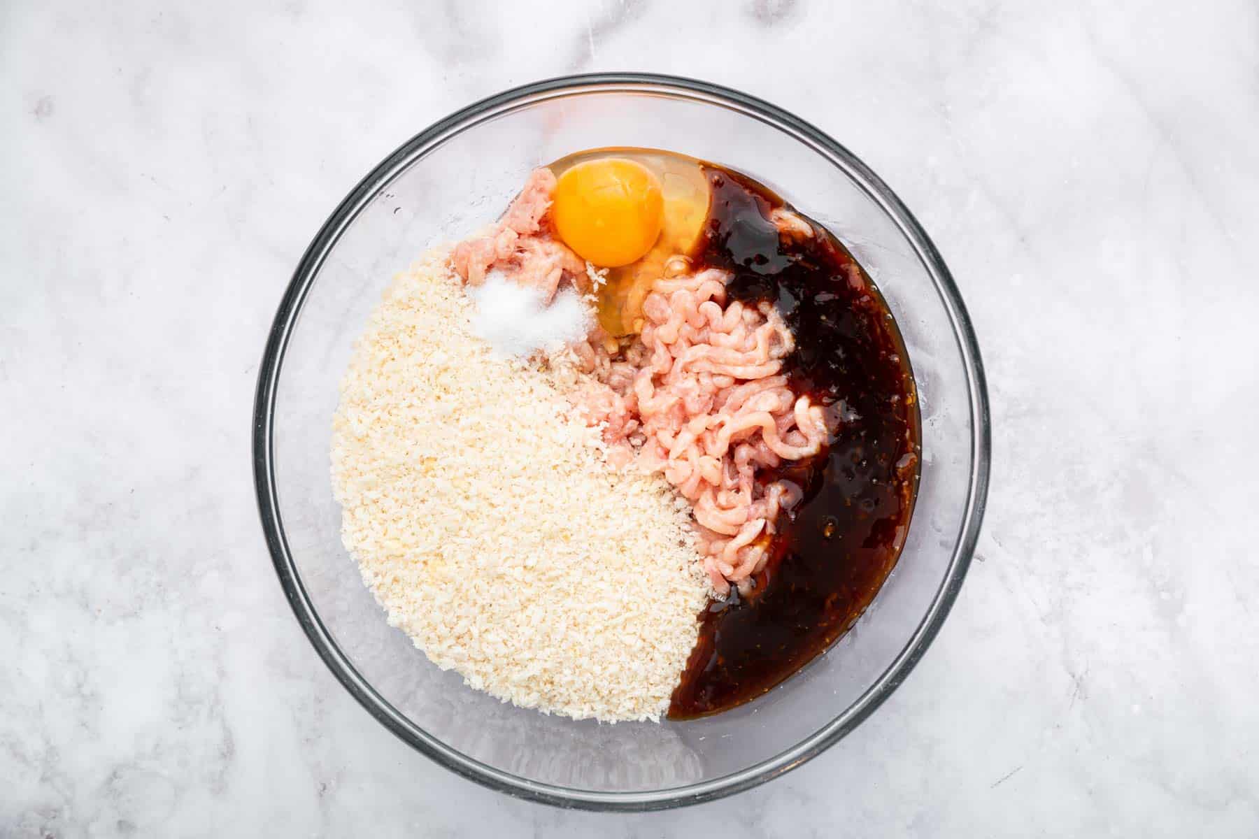 Clear bowl with ground meat, brown sauce, an egg and breadcrumbs.