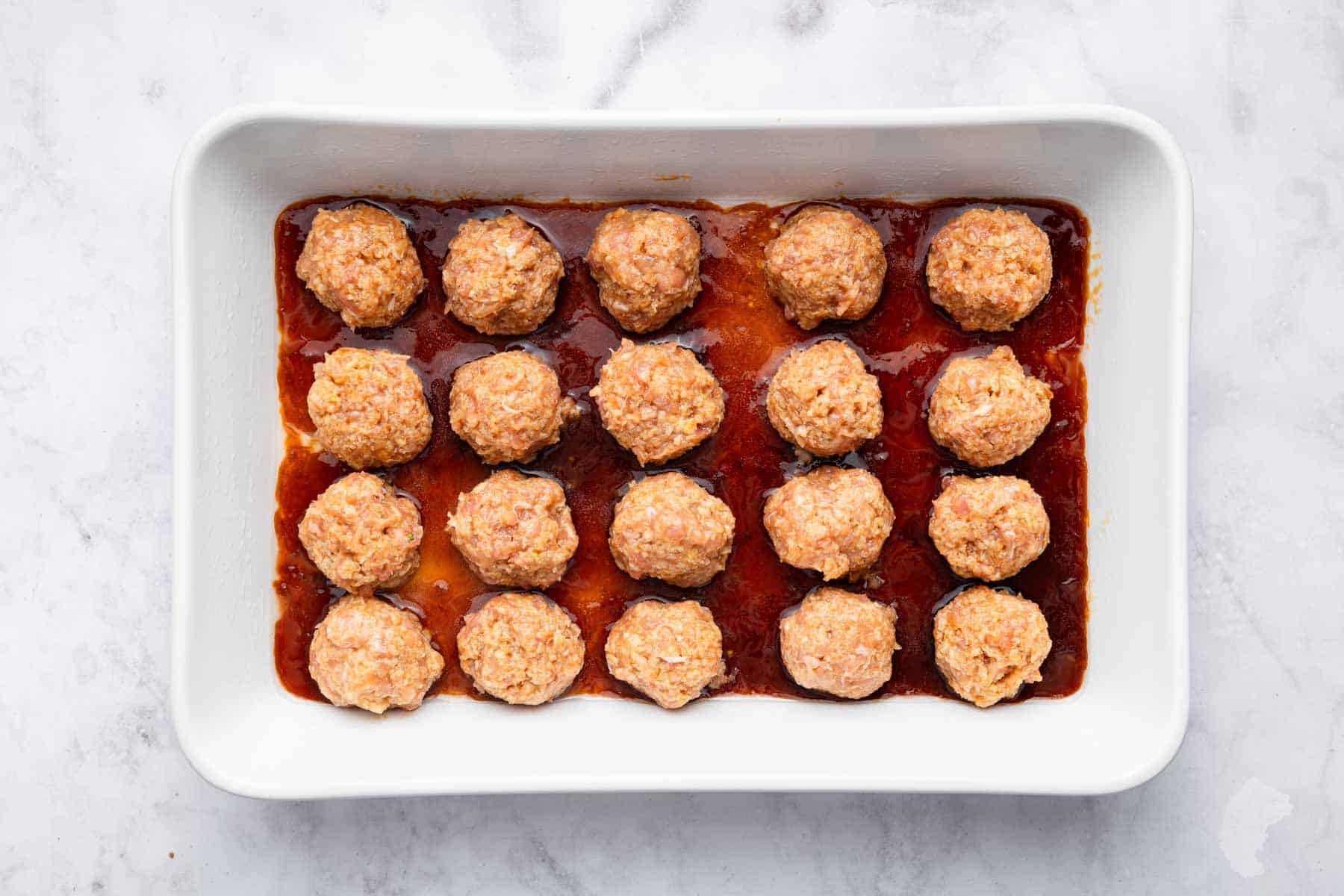 Twenty raw chicken meatballs on a bed of teriyaki sauce in a baking dish.