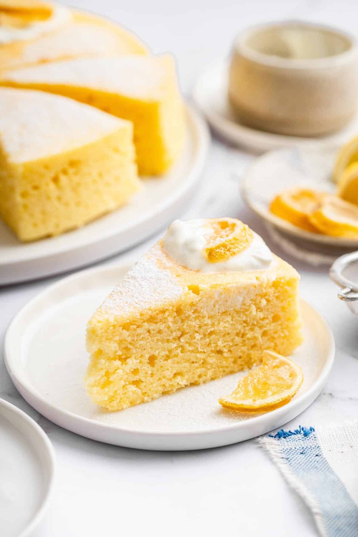 A slice of yellow yogurt cake on a white plate in front of the whole cake.