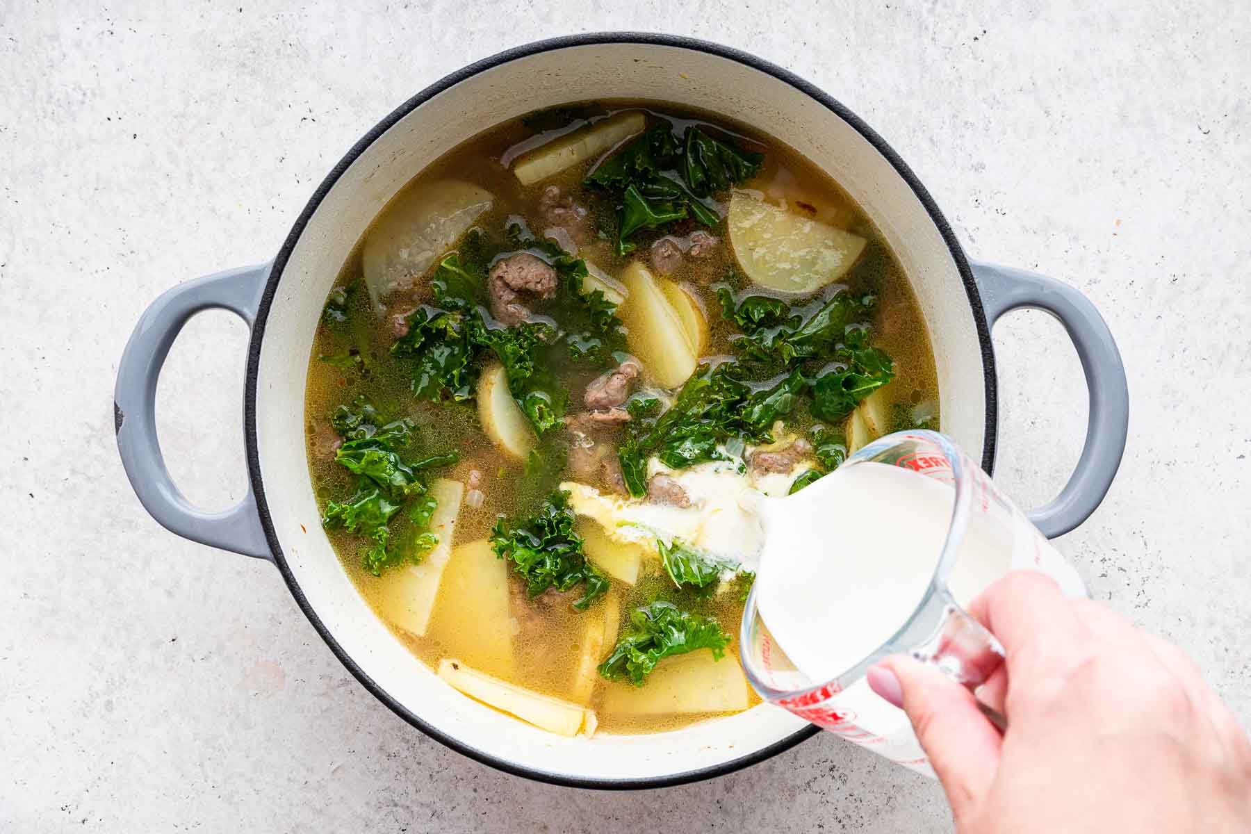 Pouring heavy cream into soup with sliced potatoes and kale.